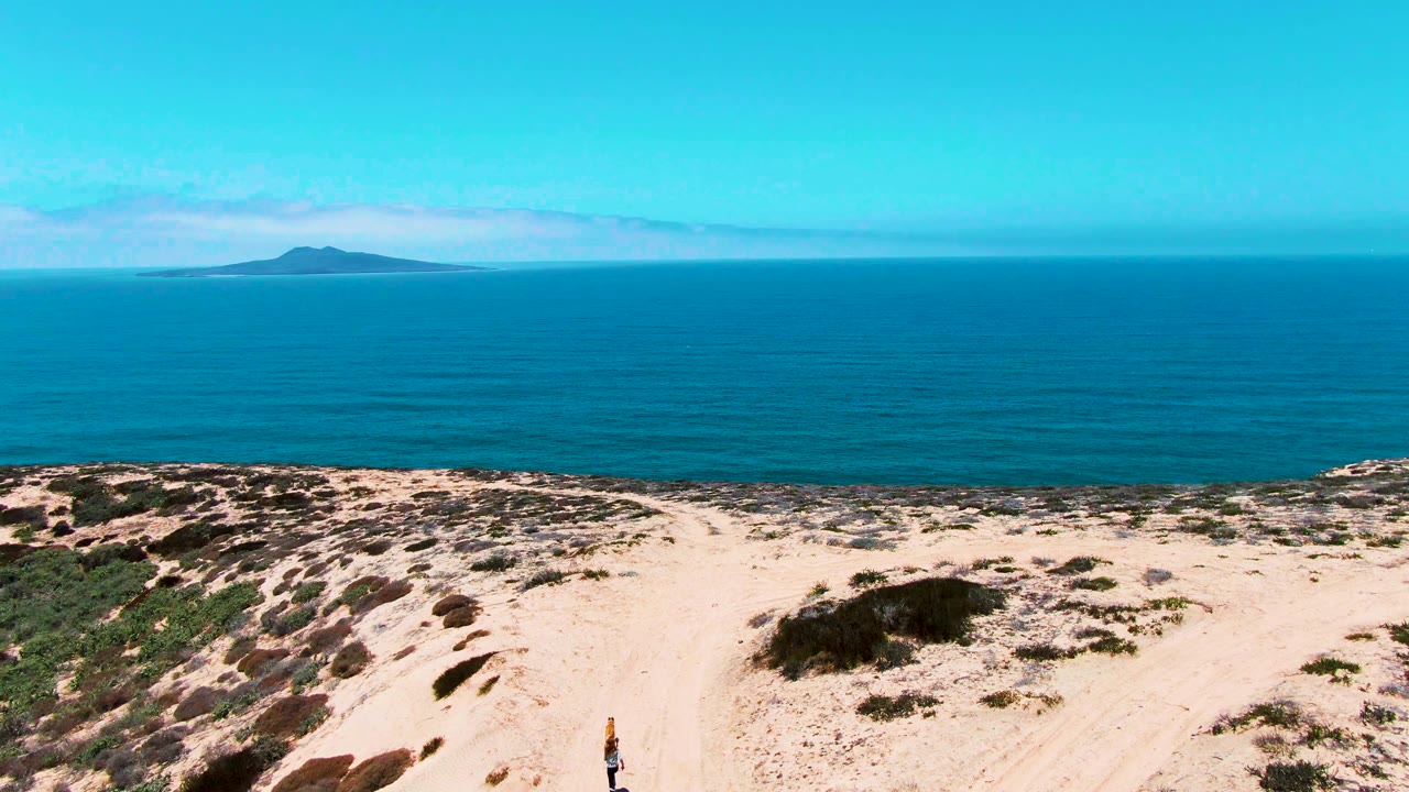 A wide San Quintin coastal dune view shows the sandy track and scrub foreground, blue ocean and island beyond, with a small person partly visible near the lower edge.