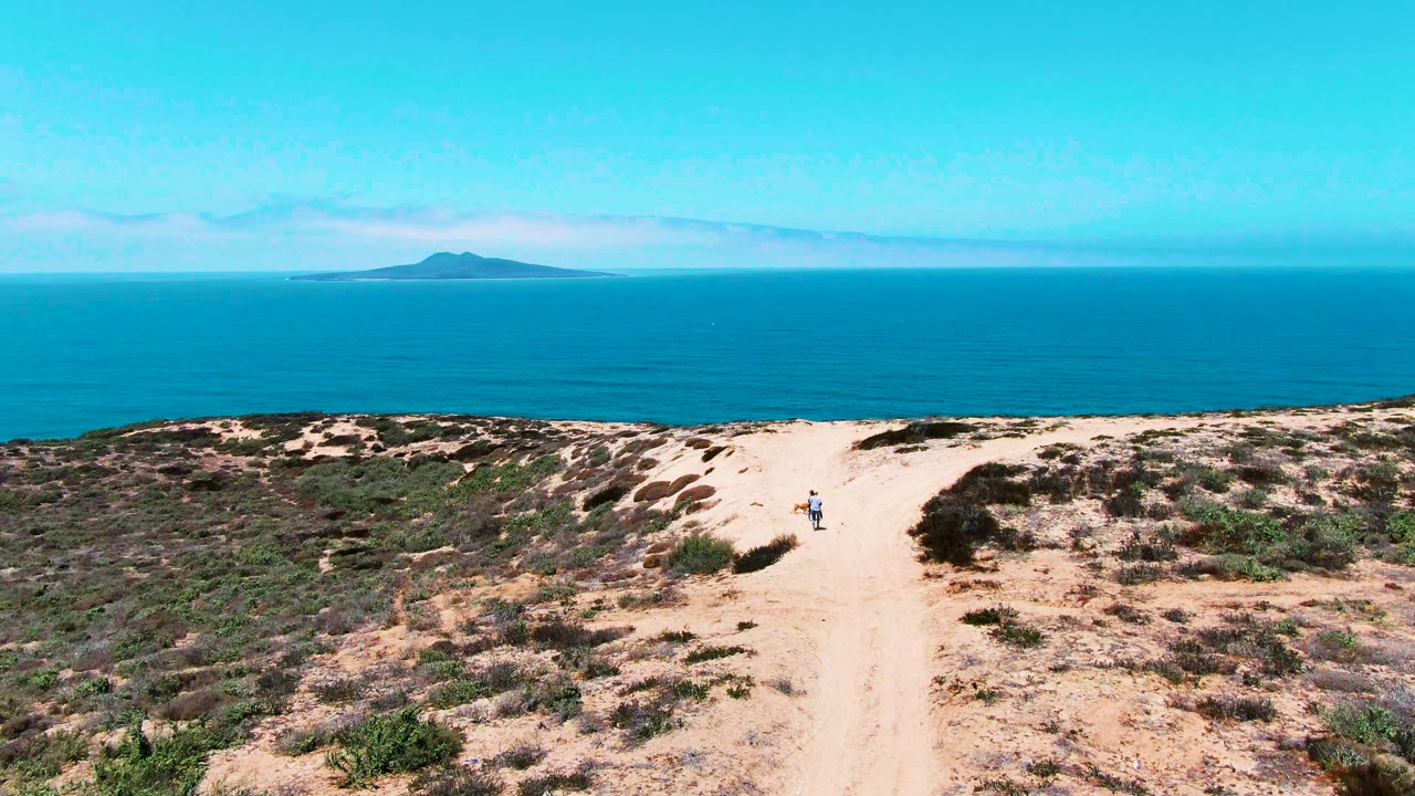 Wide elevated view in San Quintin BC Mexico of a sandy dune path leading toward blue ocean and a distant low island, with small people visible on the trail.