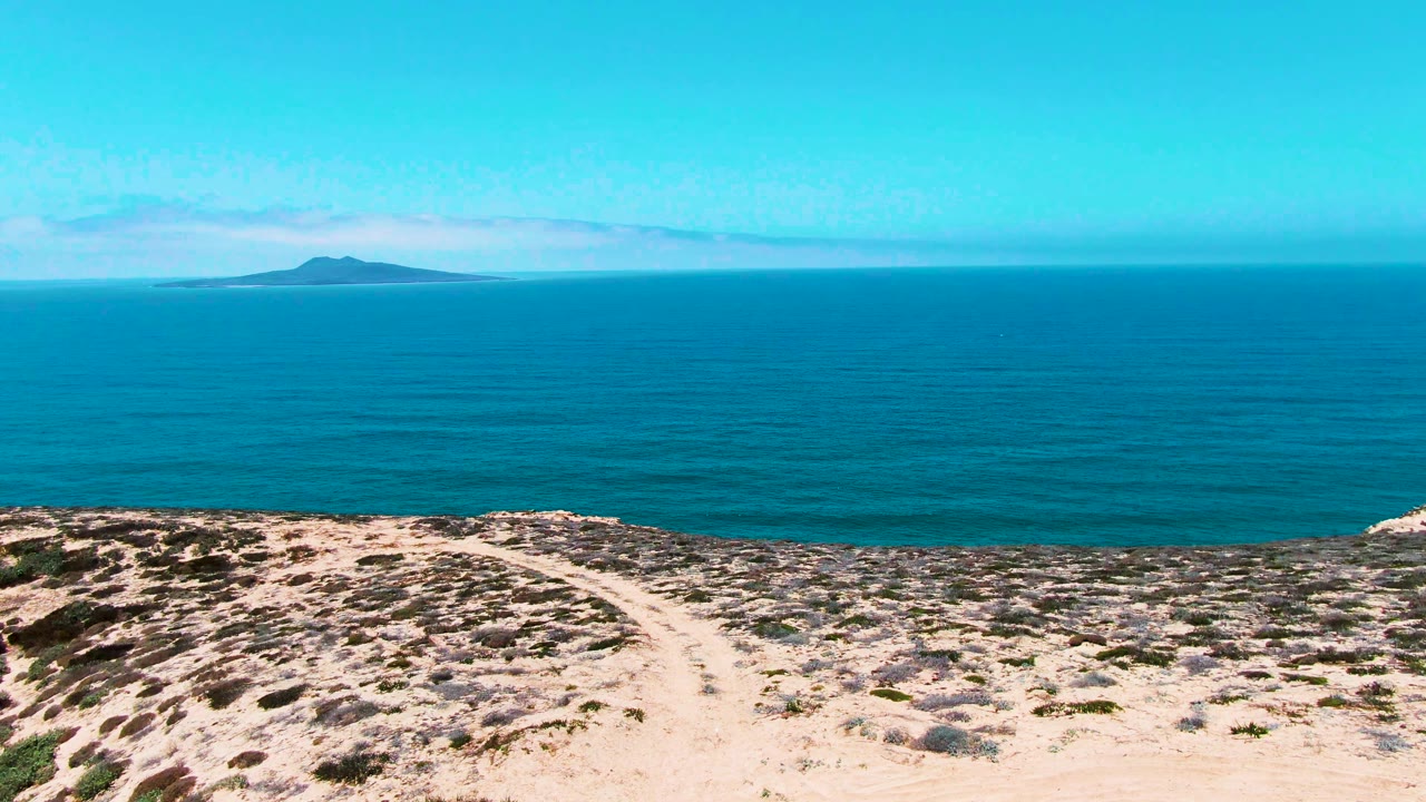 Opening frame in San Quintin BC Mexico showing a pale dirt path over scrubby rocky hillside, turquoise ocean, a low distant island on the horizon, bright sky, and no visible people.