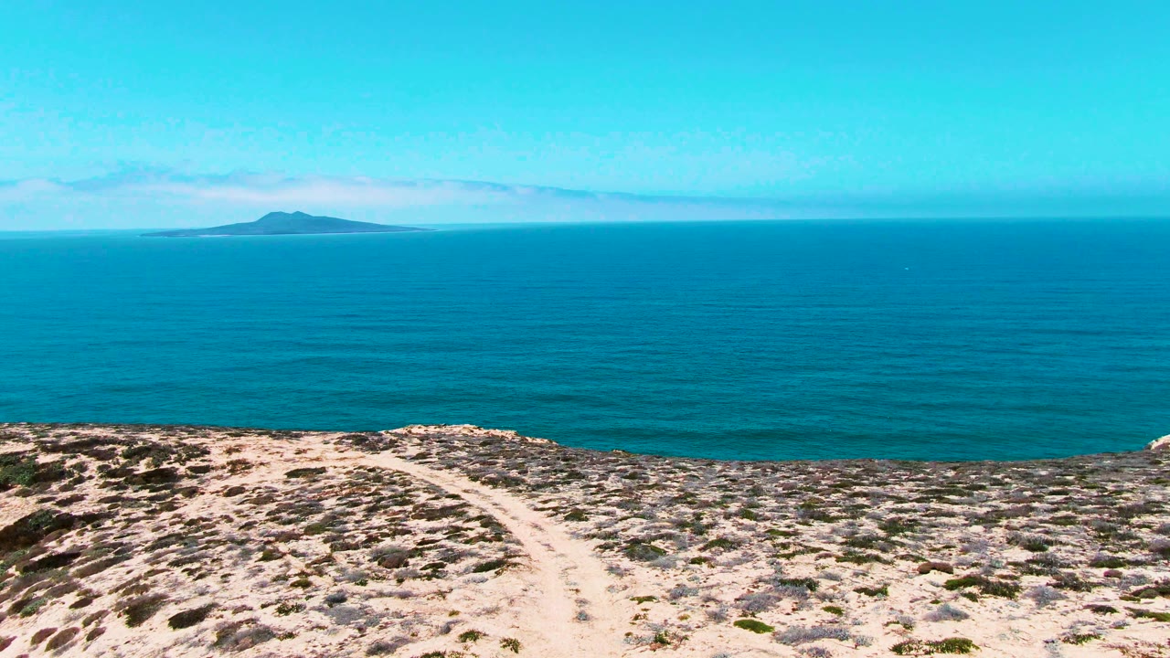 San Quintin BC Mexico frame with the rocky foreground slightly closer to the lower edge, the dirt path centered, calm blue ocean beyond, distant island left of center, and no visible people.