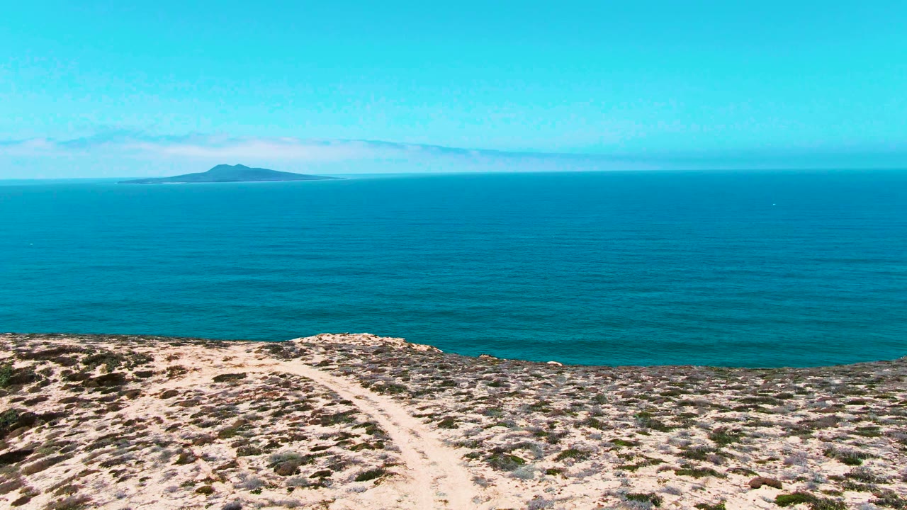 San Quintin BC Mexico view of scrub-covered arid ground and a pale footpath in the foreground, broad turquoise water across the middle, the island near the left horizon, and no visible people.