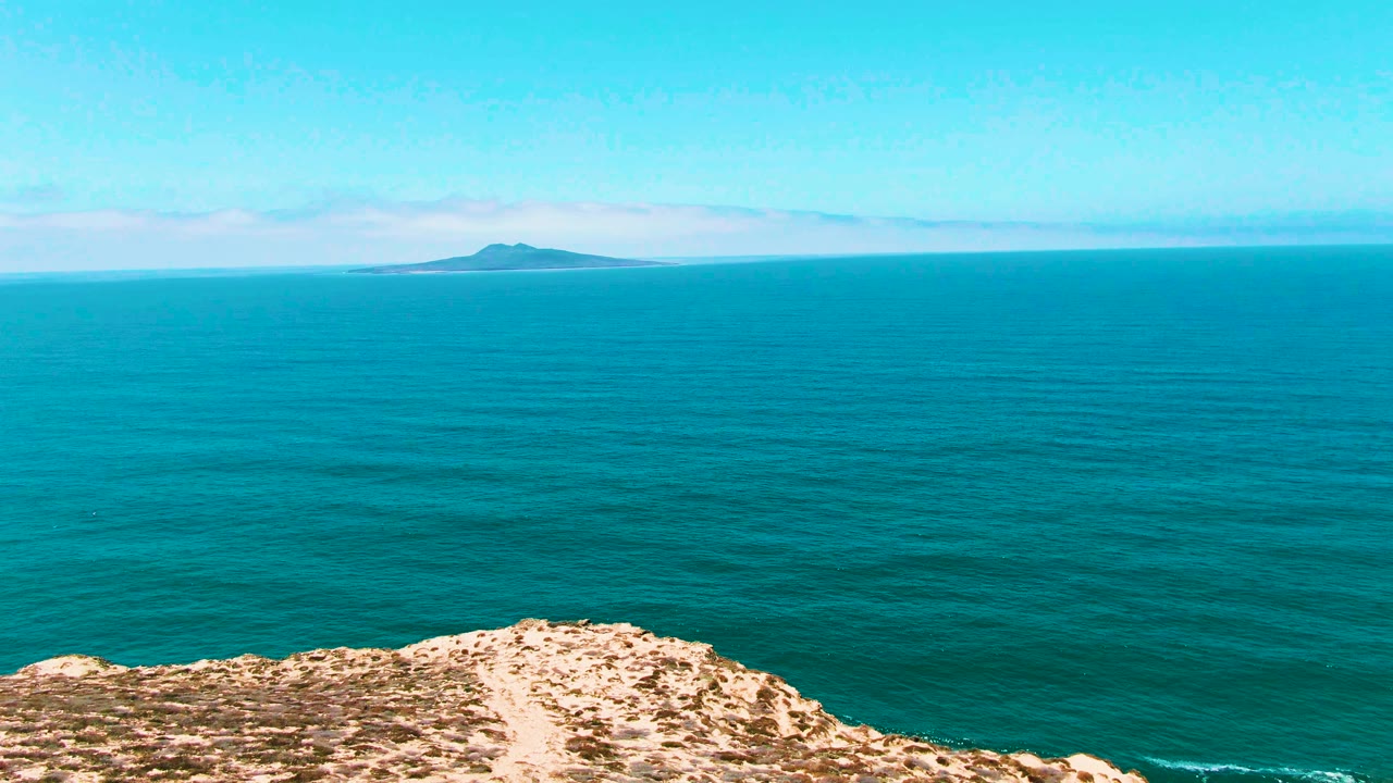 At 0:01, the San Quintin, Mexico view holds a rocky beige foreground ridge under a broad turquoise sea, with the island slightly left of center and no visible people.