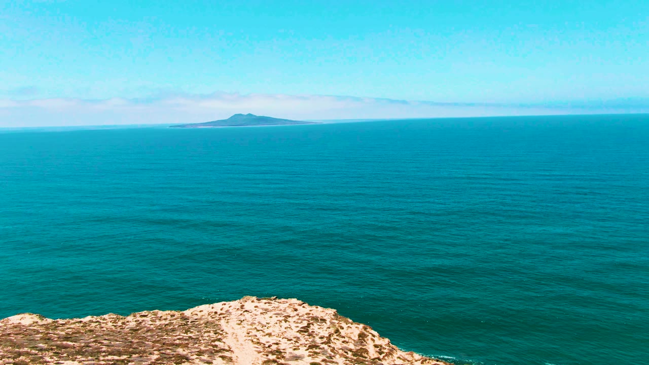 At 0:02, the San Quintin, Mexico shot shows more open water across the frame, a low rocky cliff at the bottom, a distant island near the horizon, and no visible people.