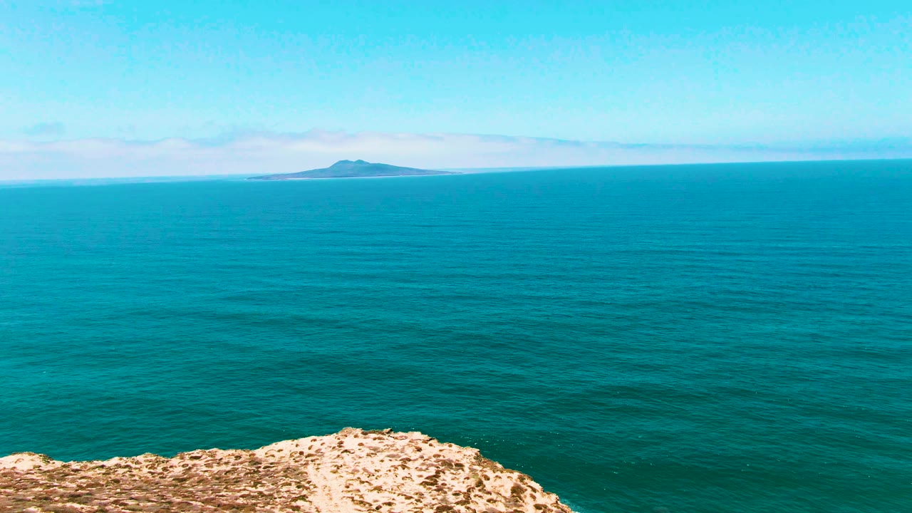 At 0:03, the San Quintin, Mexico frame shows a wide turquoise water surface, the rocky foreground dropping lower in frame, the distant island near the left horizon, and no visible people.