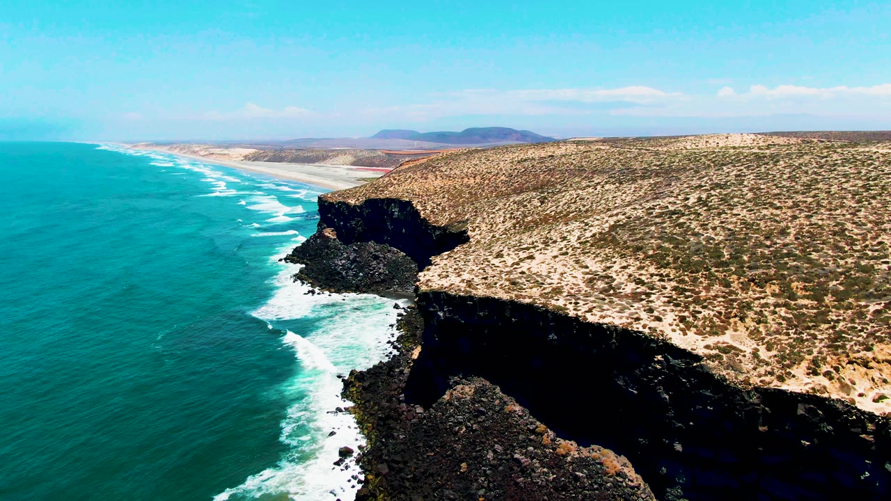 Aerial frame over San Quintin BC Mexico showing white waves curling along the base of a dark cliff, with turquoise water to the left and sparse vegetation across the plateau, no visible people.