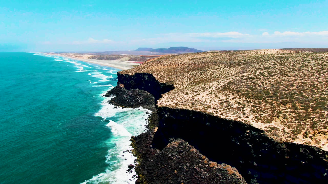 Aerial view in San Quintin BC Mexico with the rocky headland filling more of the right side, turquoise ocean stretching left, white foam tracing the shoreline, and no visible people.