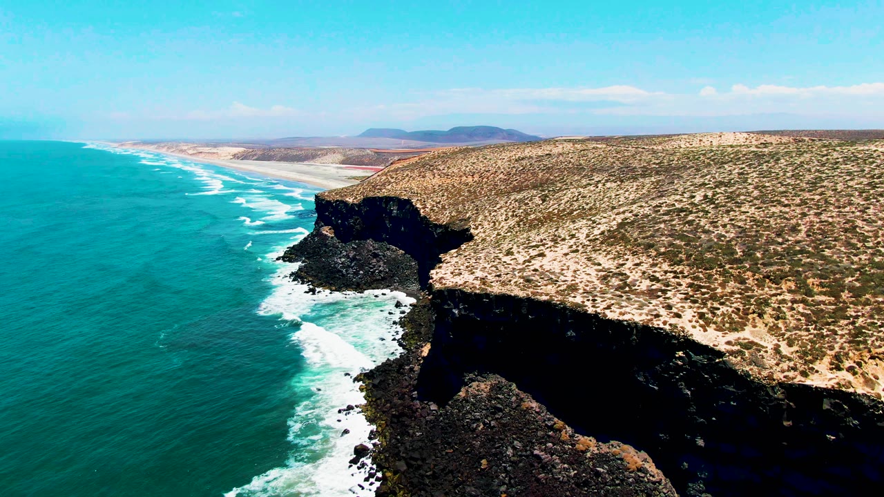 Mid-clip aerial frame in San Quintin BC Mexico looking down a long coast, with teal water and parallel surf lines to the left, a dark cliff shadow below, scrubland to the right, and no visible people.