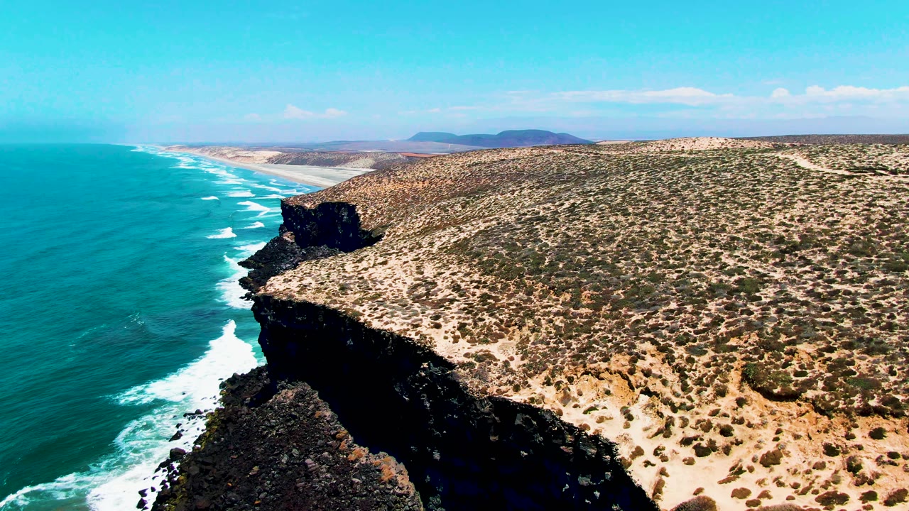 Opening frame over San Quintin BC Mexico showing turquoise ocean on the left, a dark rocky cliff in the foreground, dry scrubland inland, distant hills, and no visible people.