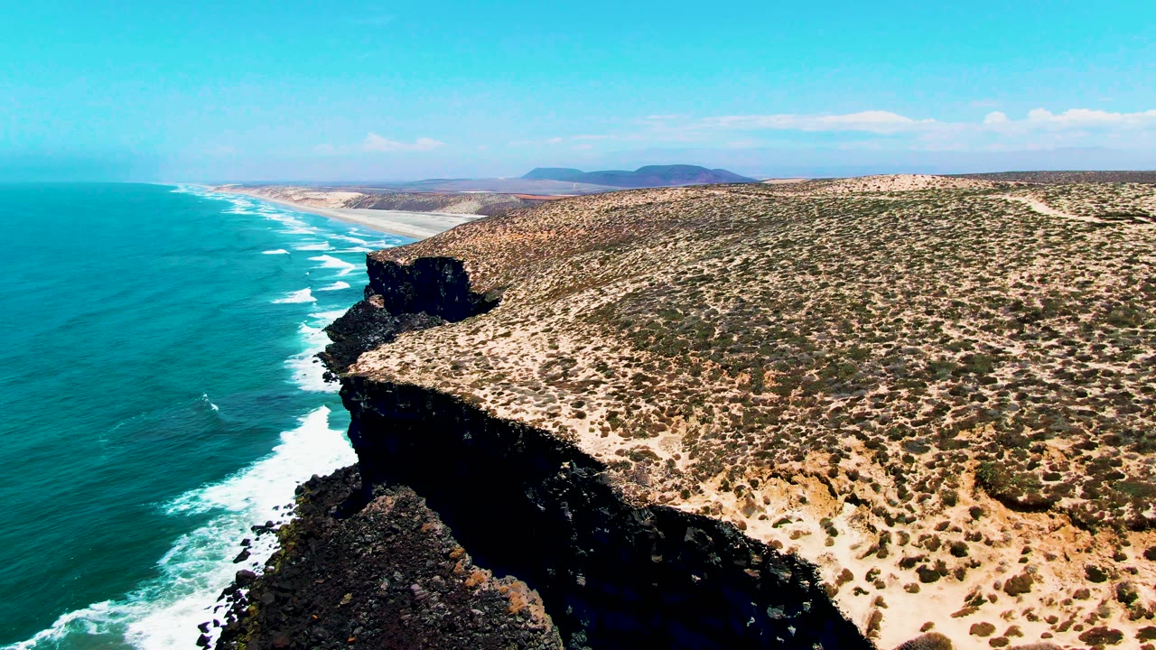 Aerial frame in San Quintin BC Mexico with the cliff face centered, white surf tracing the shoreline, scrub-covered sandy ground to the right, distant low hills, and no visible people.