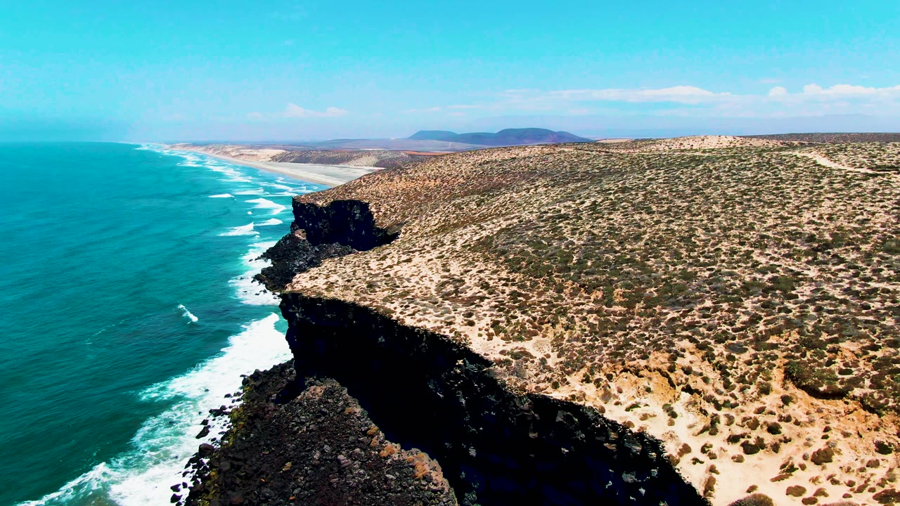Mid-clip frame over San Quintin BC Mexico showing a broad turquoise ocean expanse, a black rock shoreline below the cliff, dry brushy terrain inland, faint clouds, and no visible people.