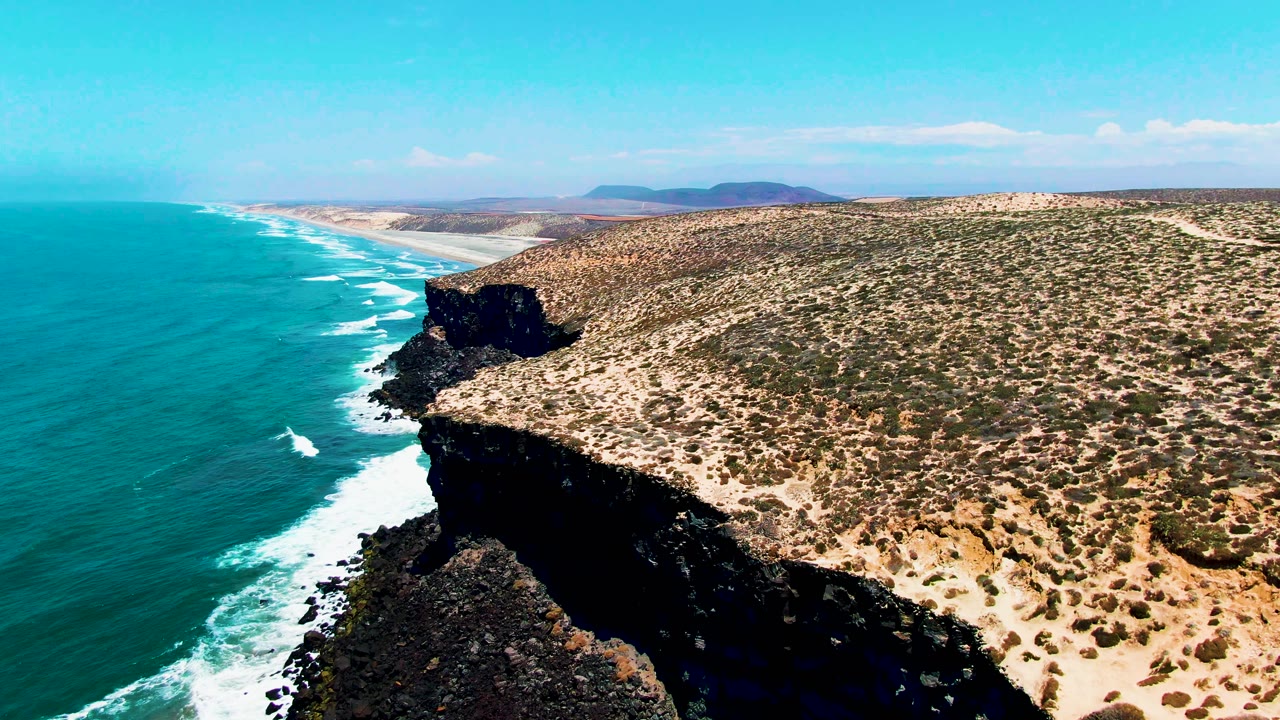 Turquoise water and surf beside dark cliffs and sandy scrubland.