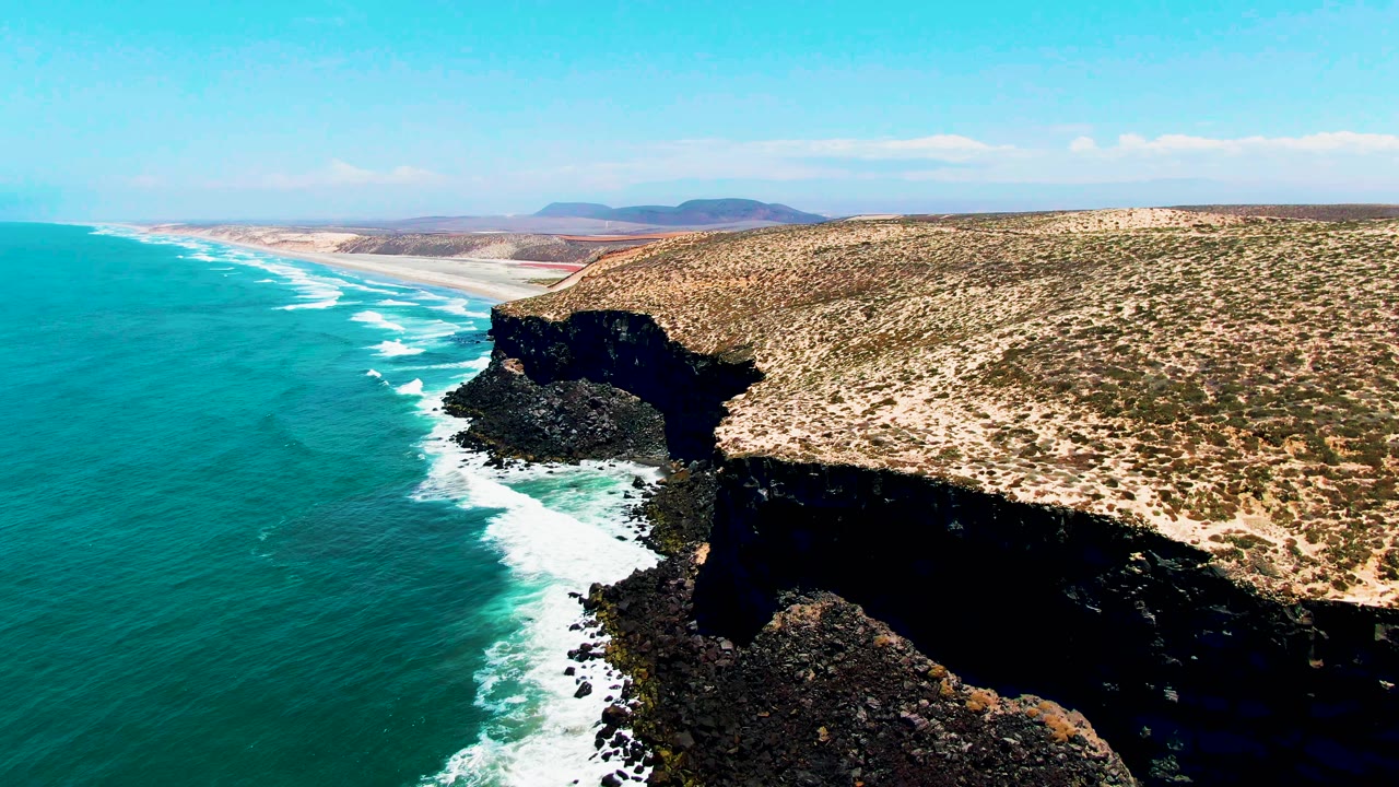 Aerial view of turquoise ocean beside dark rocky coastal cliffs and arid scrubland.