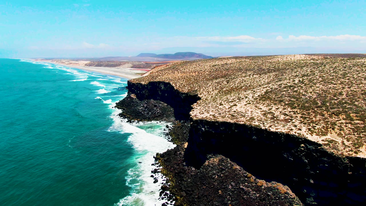 Aerial view one second into the San Quintin BC Mexico clip, with foamy surf tracing the cliff base, black rock at the shoreline, arid land above, and no visible people.