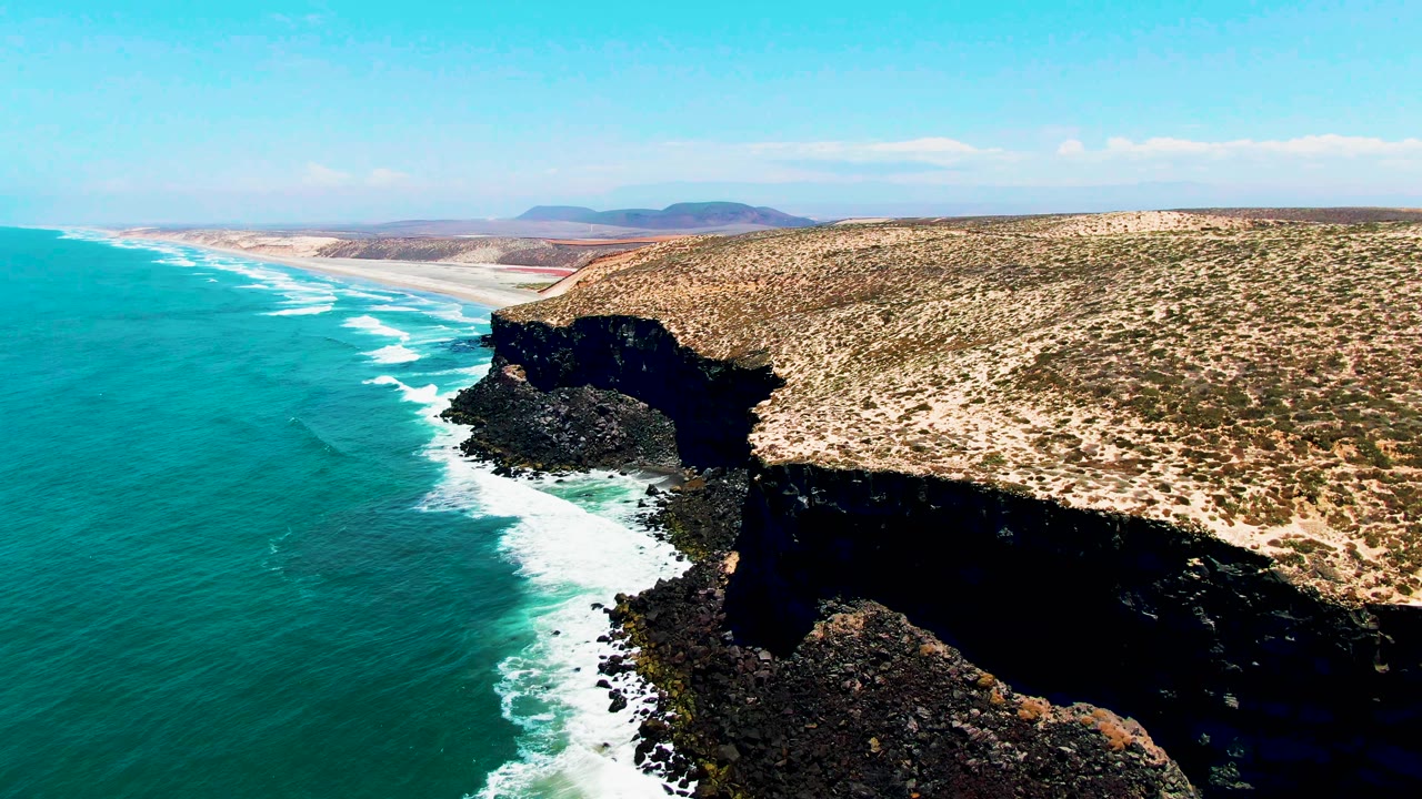 San Quintin BC Mexico aerial frame with broad blue-green ocean at left, a band of white foam below the cliff, dark rock ledges, pale scrubland inland, and no visible people.