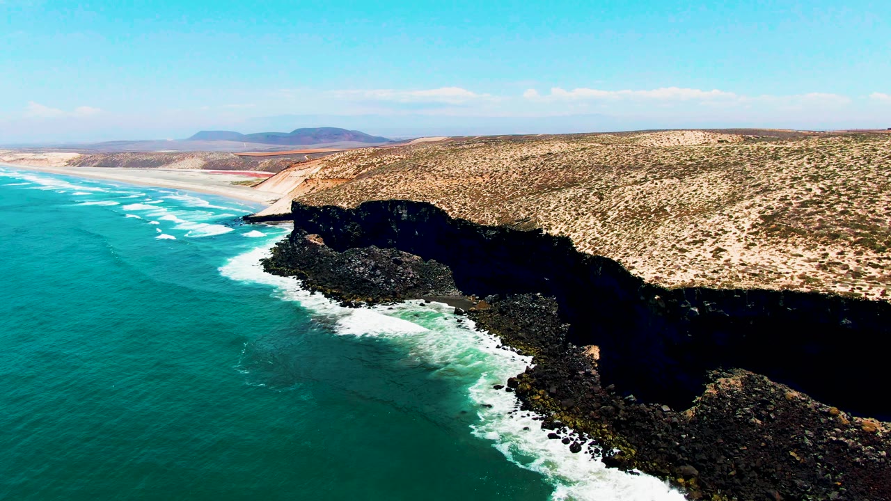 At 0:00, an aerial view over San Quintin, BC Mexico shows turquoise water on the left, white surf on dark rocks, a sandy bluff to the right, distant hills, and no visible people.