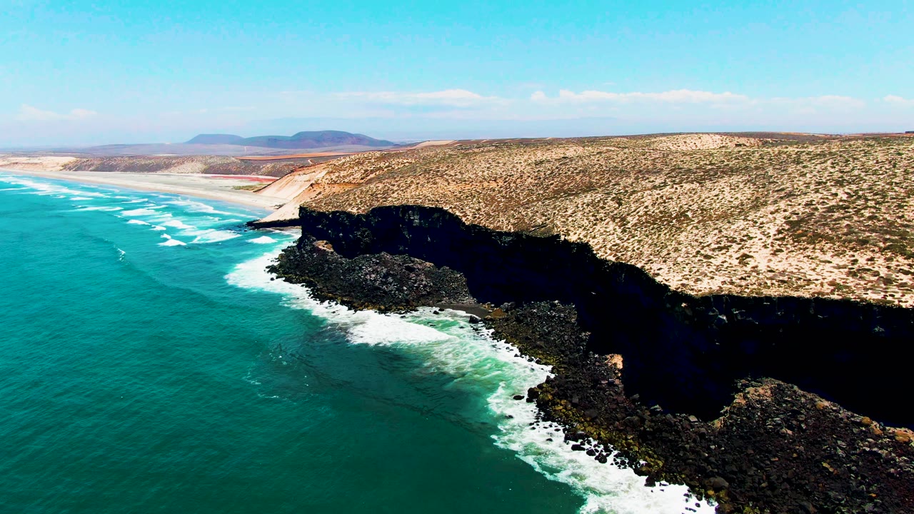 Turquoise waves roll toward a dark rocky shoreline below a sandy bluff and clear sky.