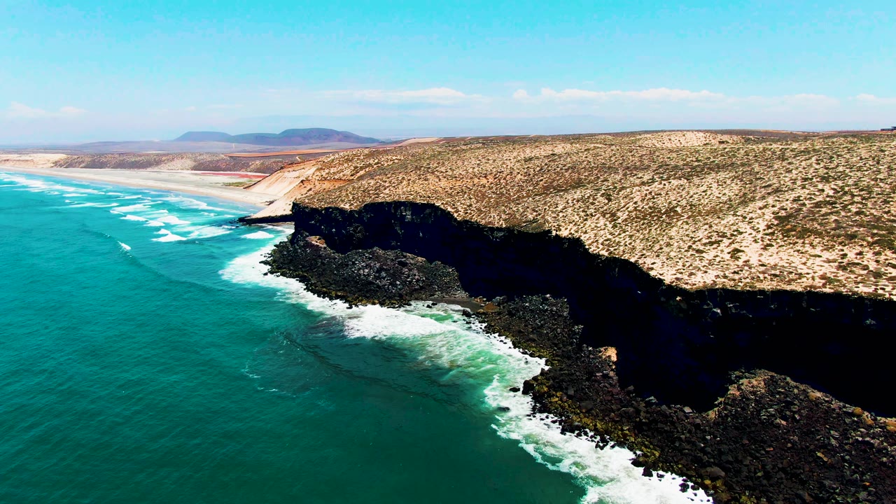 At 0:02, the aerial angle over San Quintin shows a longer run of pale beach in the background, surf curling near the rocky point, and no visible people.