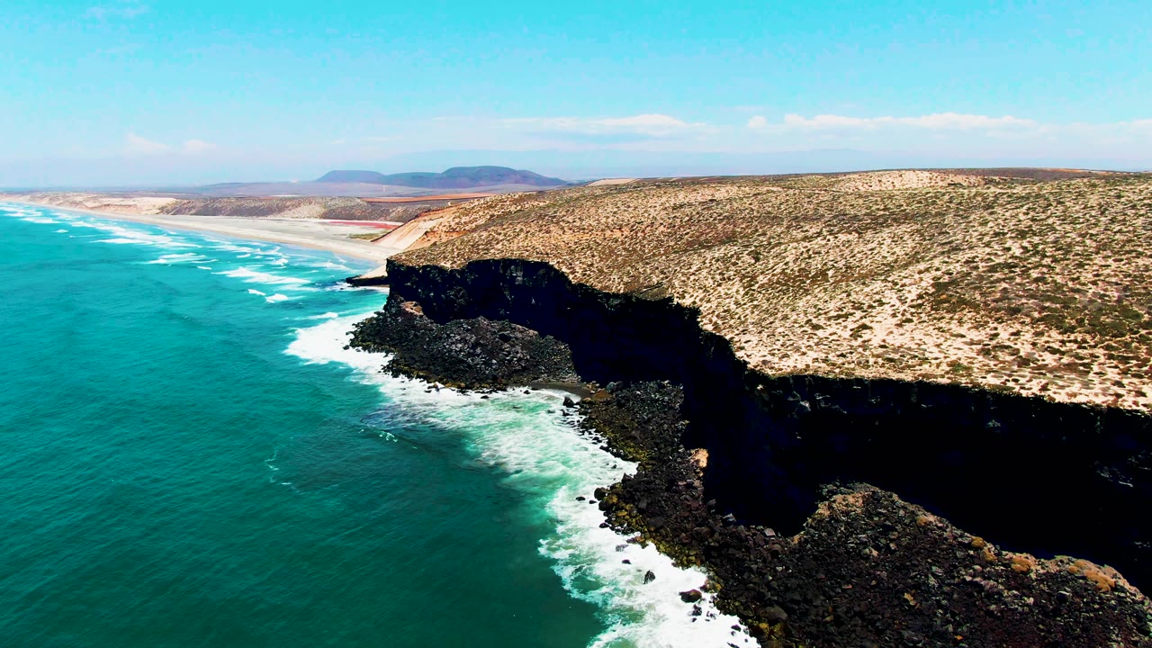 Aerial frame from San Quintin BC Mexico showing turquoise water on the left, white surf around dark rocky cliffs, pale beach beyond, distant hills, bright sky, and no visible people.