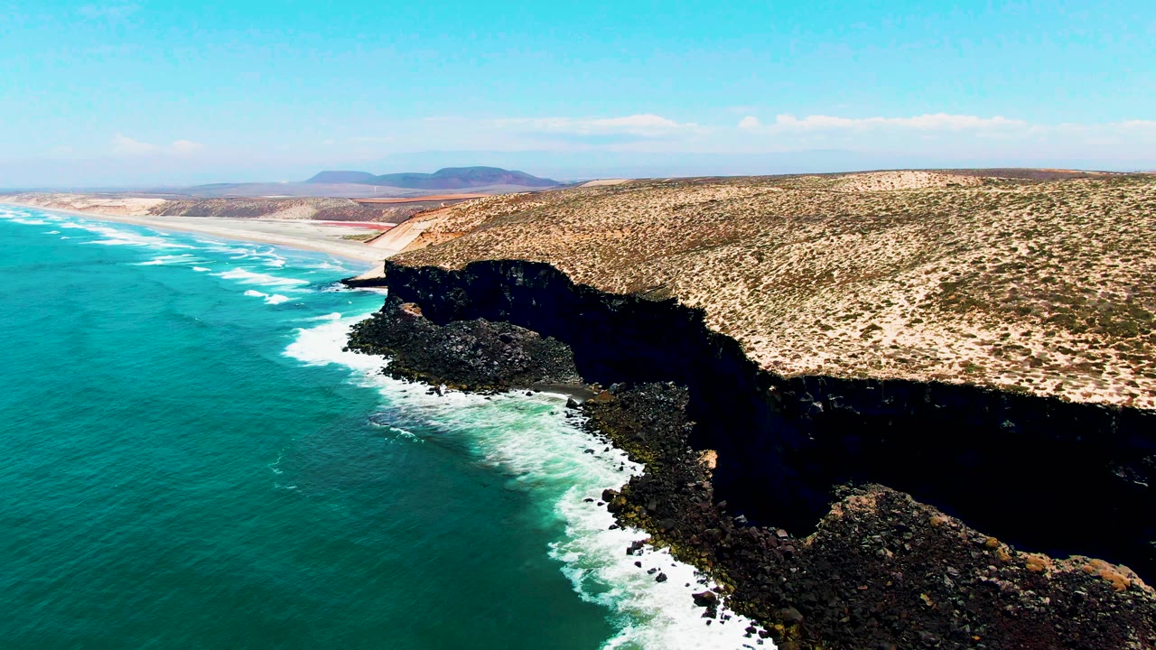 Aerial view in San Quintin BC Mexico with white foam tracing the cliff base, turquoise sea filling the left half, sandy shoreline receding behind the headland, and no visible people.