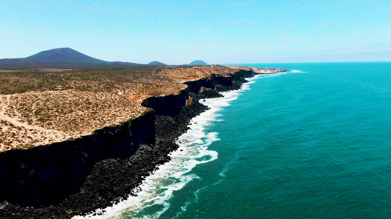 Aerial frame from San Quintin, Mexico, showing dry scrubland above dark rocky cliffs and turquoise ocean with white surf along the shore, no visible people.
