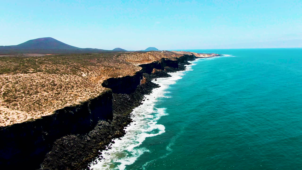 Aerial view in San Quintin, Mexico, where white surf bands trace the rocky cliff base while open turquoise water fills the right side, with no visible people.