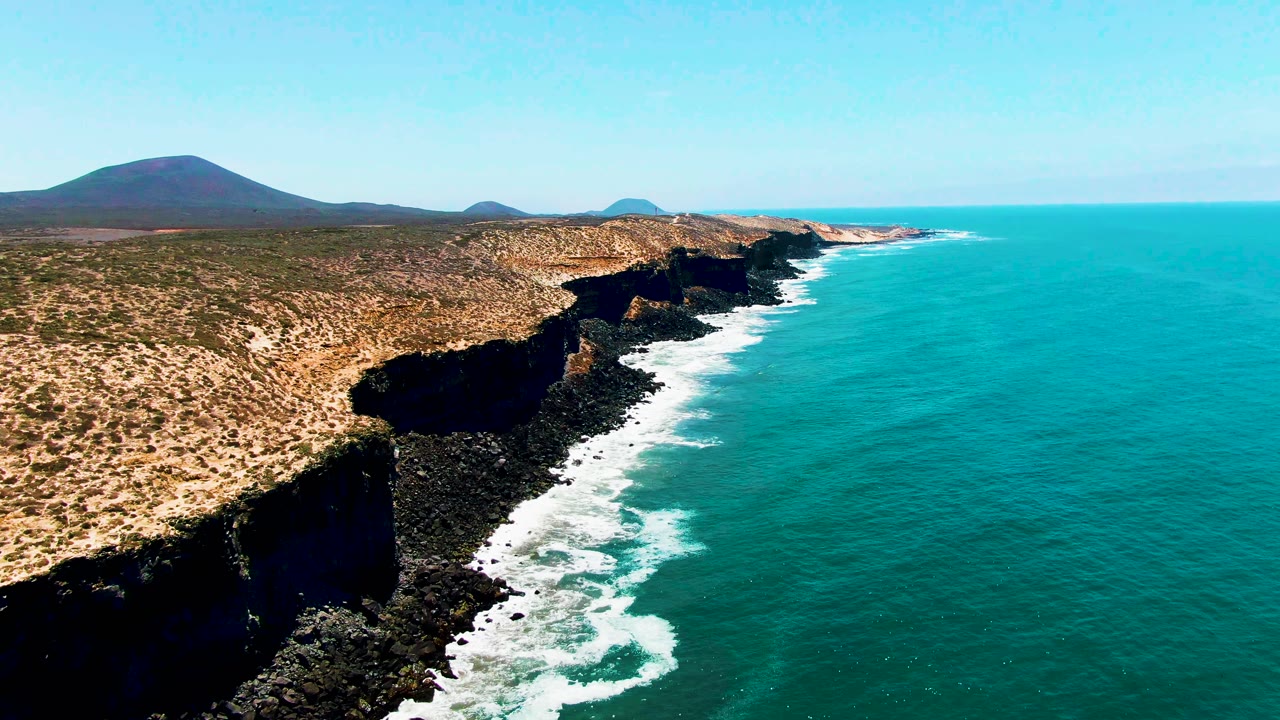 Aerial view in San Quintin BC Mexico of black rocky cliffs meeting turquoise ocean, with dry scrubland and low hills in the background and no visible people.