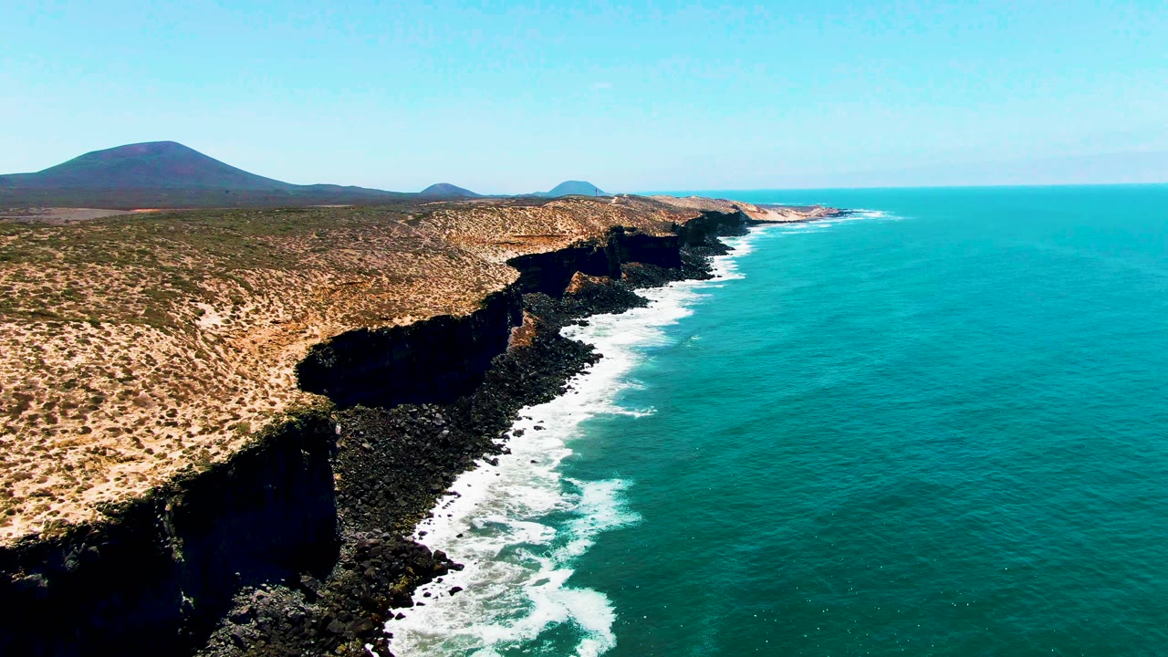 Aerial frame over San Quintin BC Mexico shows white waves curling along dark rocks, dry tan land above the cliffs, distant hills, and no visible people.
