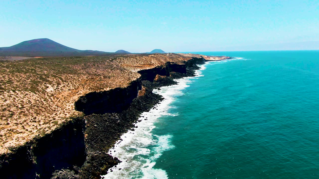 San Quintin BC Mexico coastline appears from above with dry scrubland on the left, a dark cliff notch near the shore, turquoise water to the right, and no visible people.