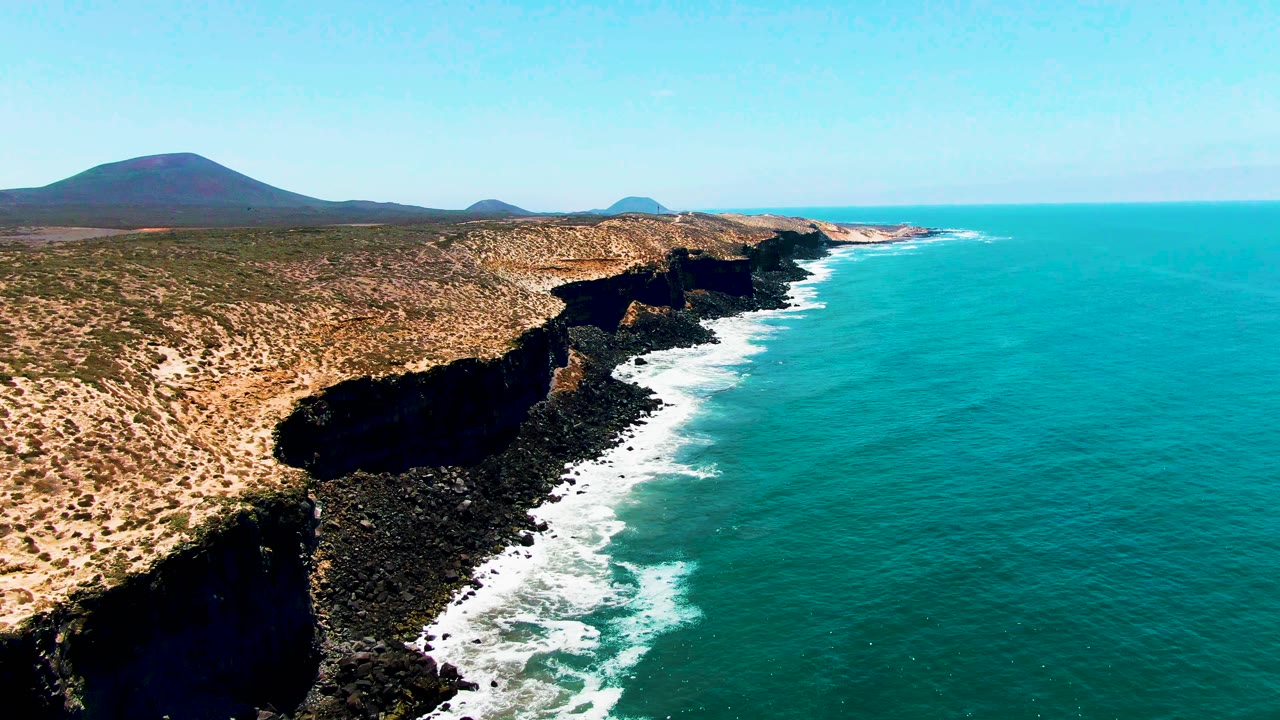 Coastline of rocky cliffs with surf and distant hills under blue sky.