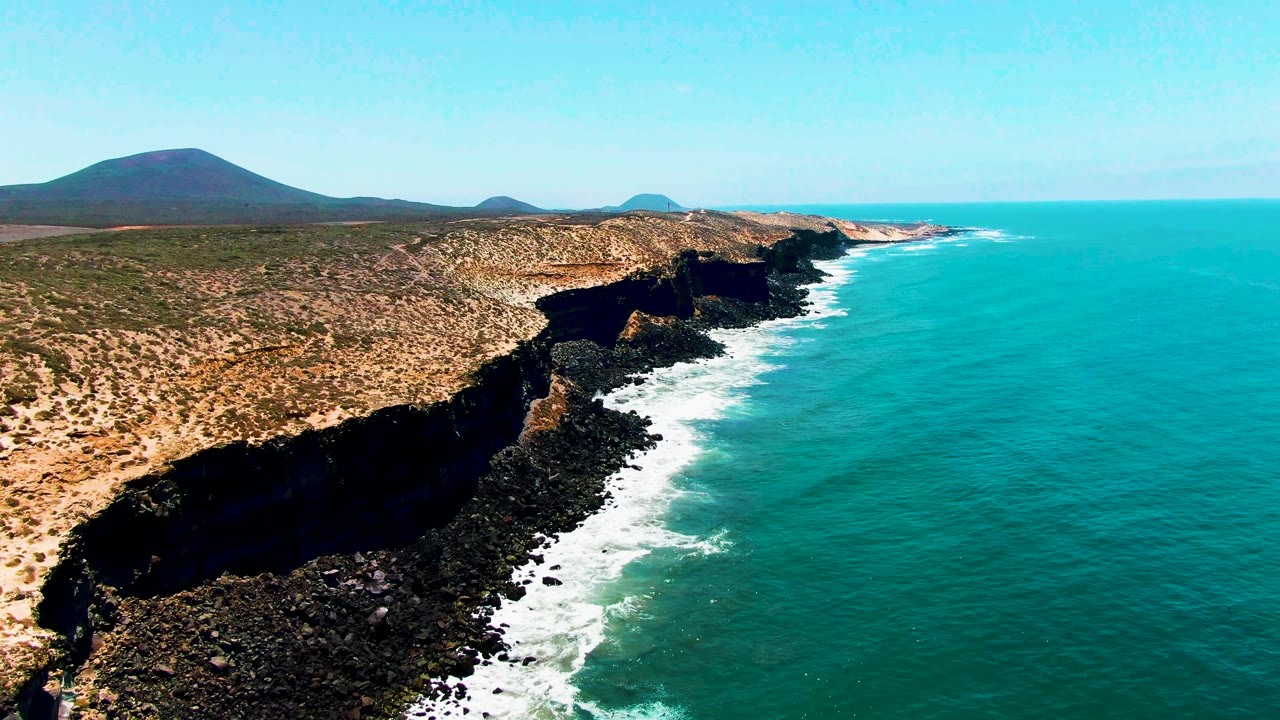 Aerial frame over the San Quintin, Mexico coast showing dark rocky cliffs, white surf, turquoise water, dry scrubland, distant hills, and no visible people.