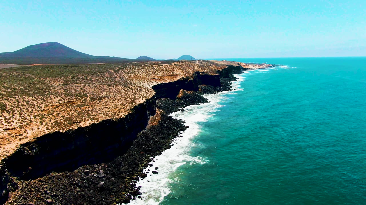 Aerial view along the San Quintin, Mexico shoreline with foamy waves tracing the black rocks, scrub-covered land inland, low hills on the horizon, and no visible people.