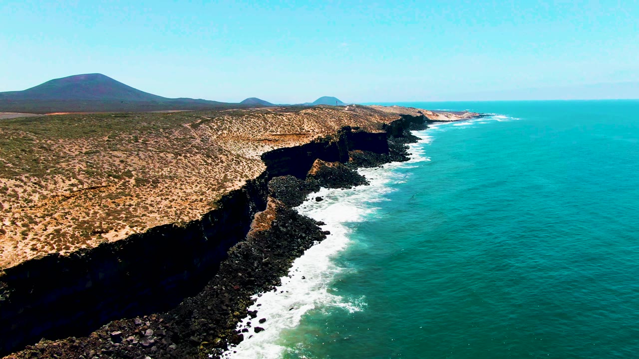 Aerial coastline with dark cliffs, surf, and distant rounded hills.
