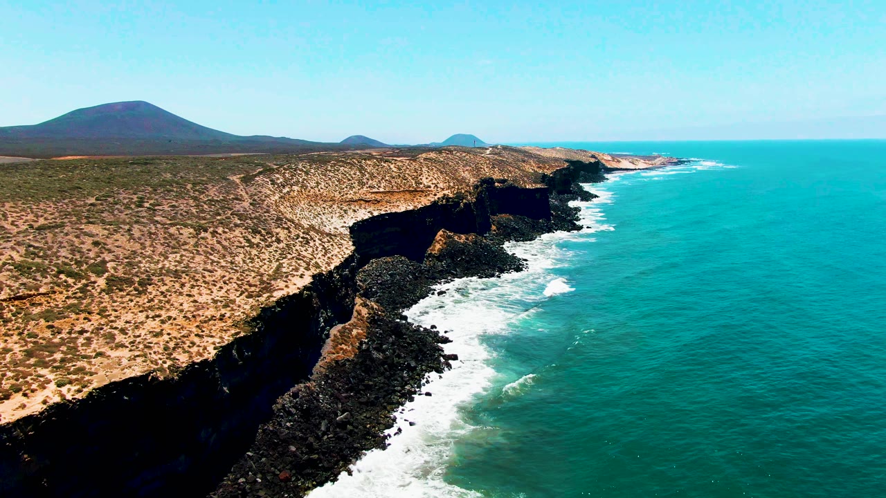 Aerial view along the San Quintin BC Mexico shore with a broad dry bluff, black cliff edges, foaming waves at the base, open turquoise water, distant hills, and no visible people.