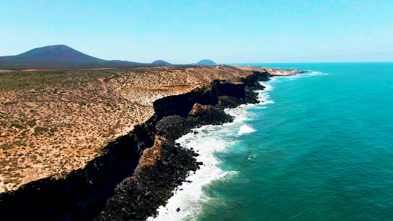 High aerial frame of the San Quintin BC Mexico coastline with jagged dark cliffs running diagonally through the image, turquoise water filling the right side, pale dry terrain above, and no visible people.