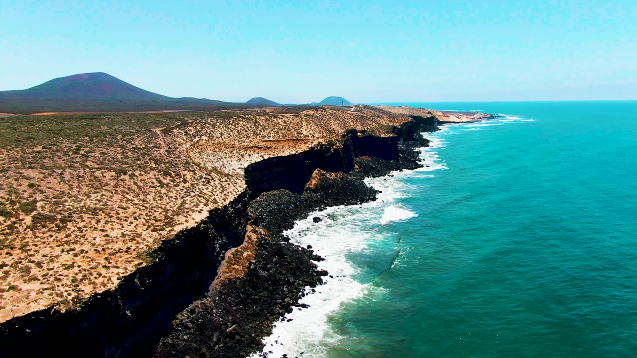 San Quintin BC Mexico aerial frame with the coastline set farther left, waves curling against dark rocks below the cliff, desert-like land stretching inland, blue ocean to the right, and no visible people.