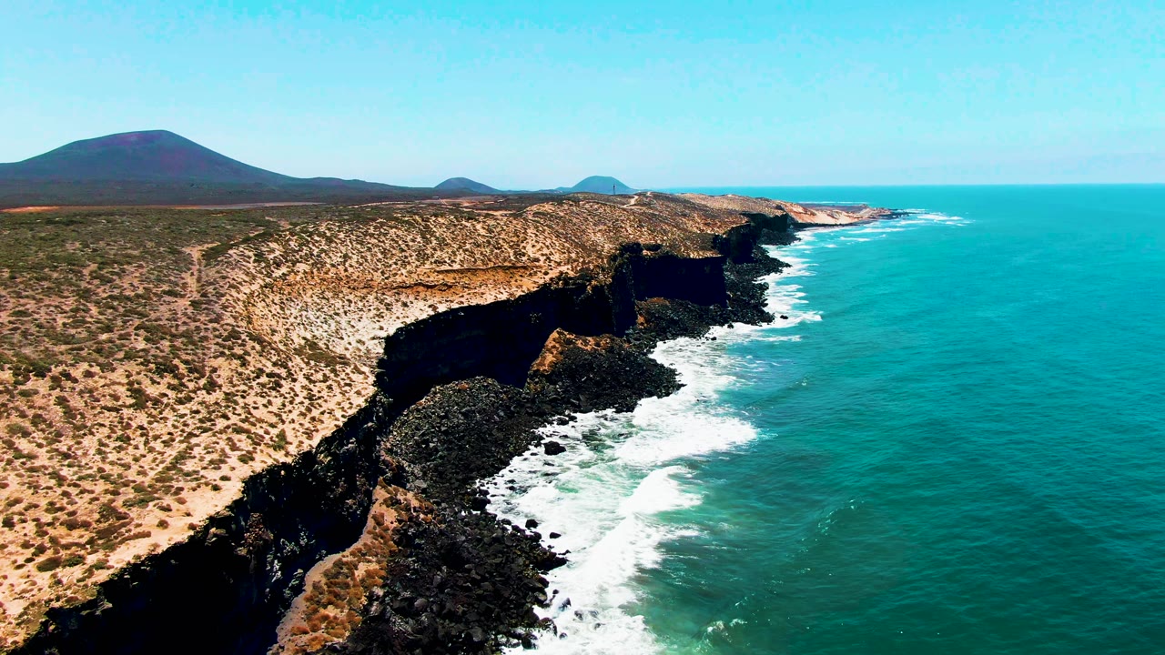 Aerial frame from San Quintin BC Mexico showing turquoise water on the right, dark rocky cliffs along the shore, tan scrubland on the left, distant hills on the horizon, and no visible people.