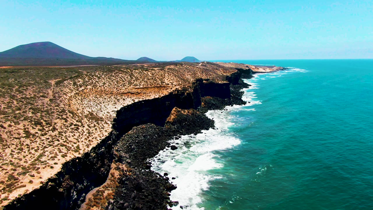 Aerial view in San Quintin BC Mexico with the cliff edge slightly more centered, white foam running along black rocks, scrub-covered tan ground inland, blue ocean to the right, and no visible people.