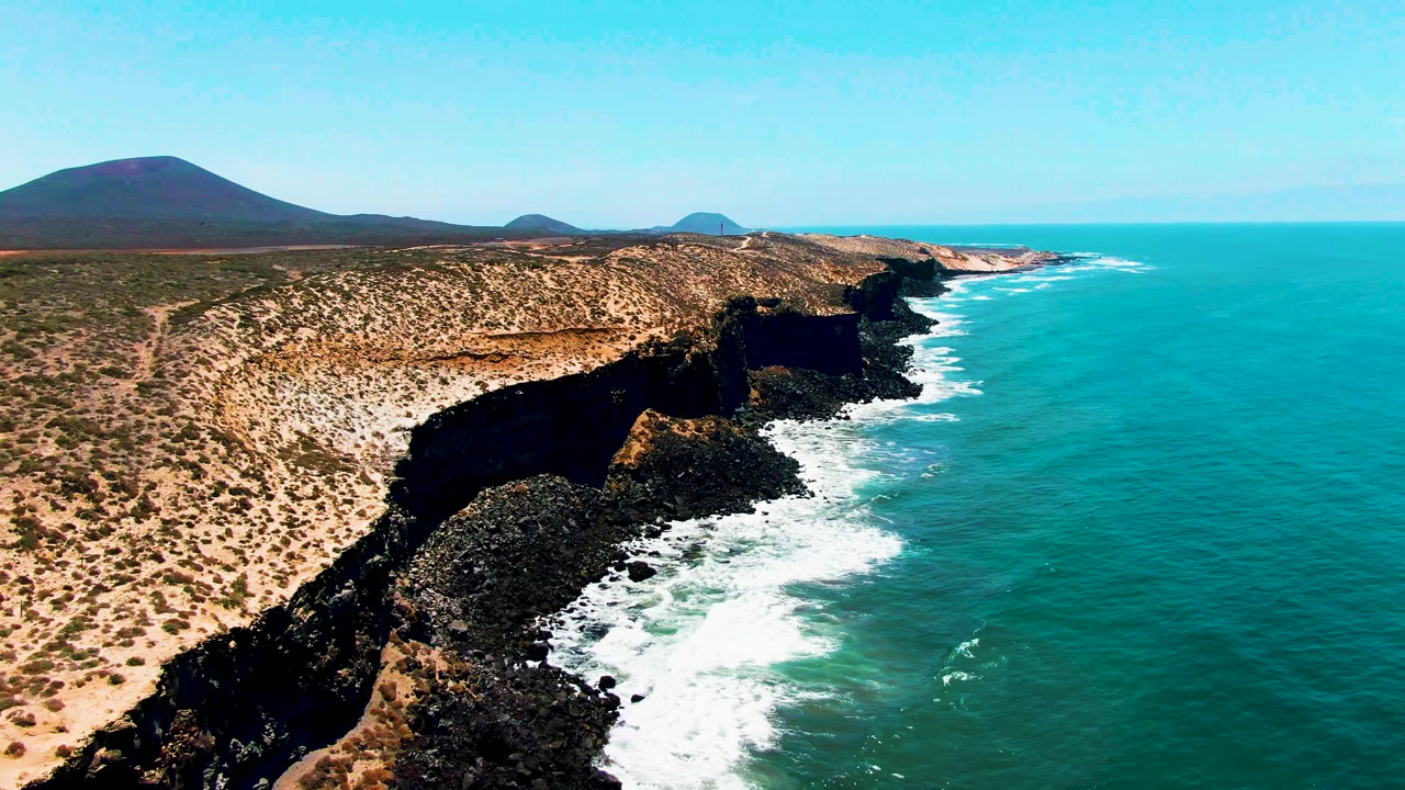 San Quintin BC Mexico aerial frame showing a longer stretch of rocky coast, bright white surf spreading over the near shoreline, open turquoise sea, dry vegetated land, distant hills, and no visible people.