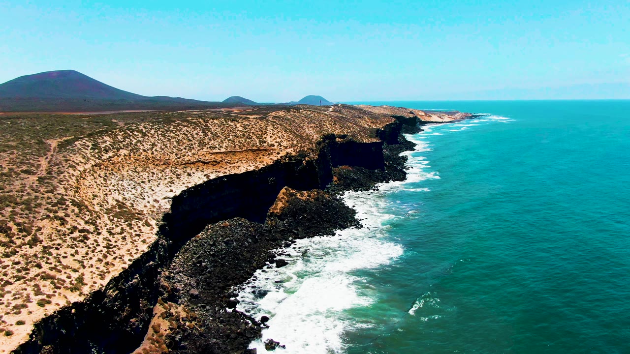 Wide aerial view of arid coastline, cliff shadows, and waves along the shore.
