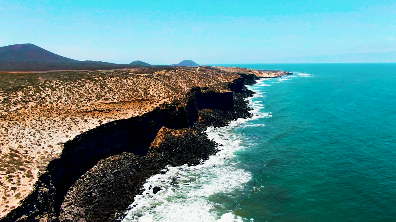 Aerial frame over San Quintin cliffs, showing black rock at the waterline, white surf curling into the shore, dry scrubland above, and no visible people.