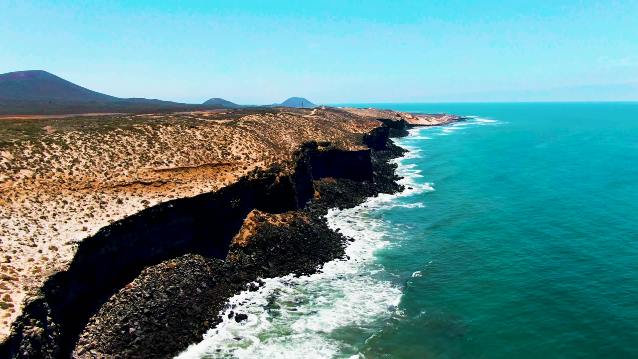 San Quintin coastal aerial with a desert plateau stretching across the left side, a jagged dark cliff face below, bright turquoise sea on the right, and no visible people.