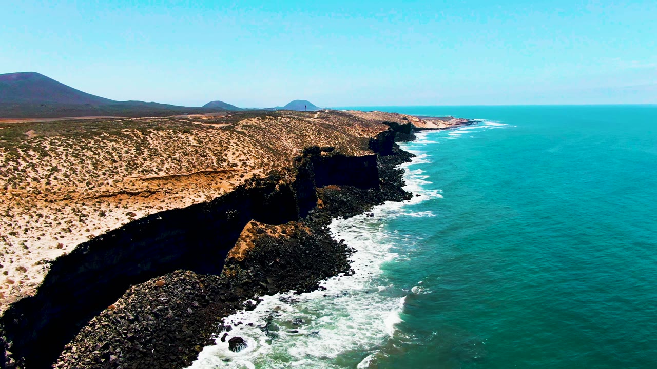 Aerial view of San Quintin coastline with the nearest dark cliff dropping into foamy water, tan scrubland continuing inland, rounded hills on the horizon, and no visible people.
