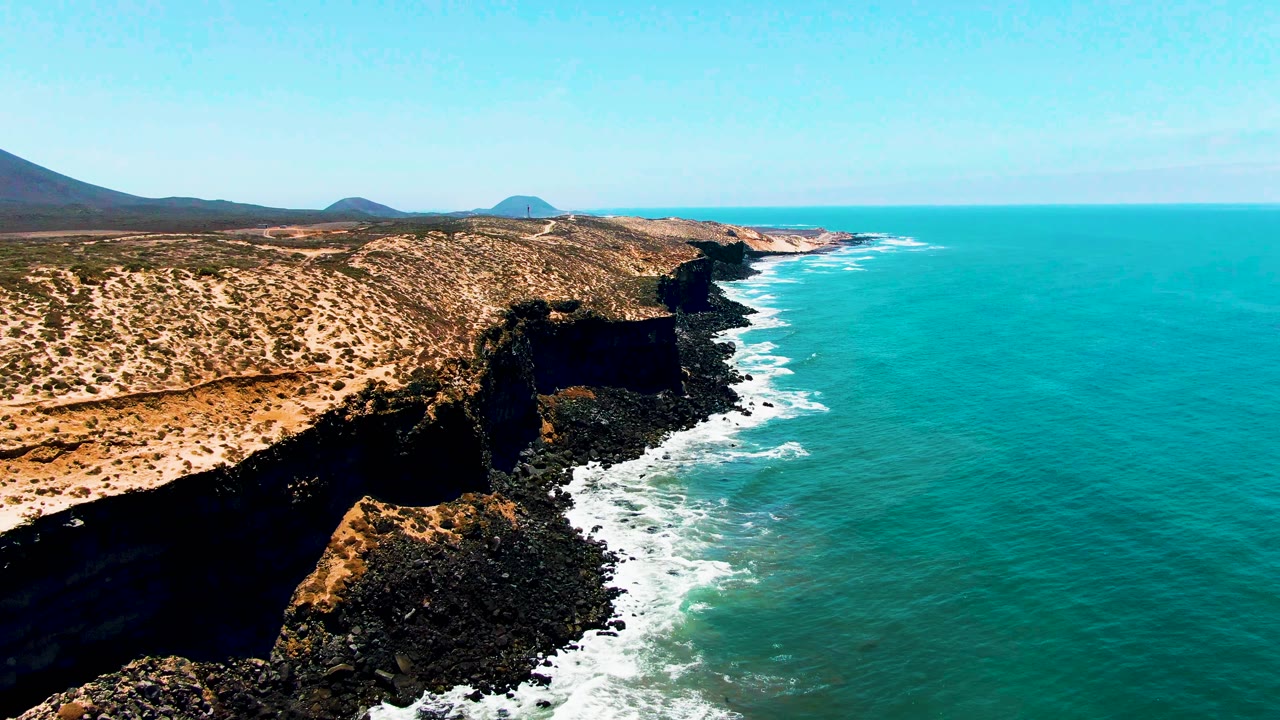 San Quintin BC Mexico coastline with dark cliffs, tan arid terrain, turquoise water, white surf, distant hills, and no visible people at the start of the clip.