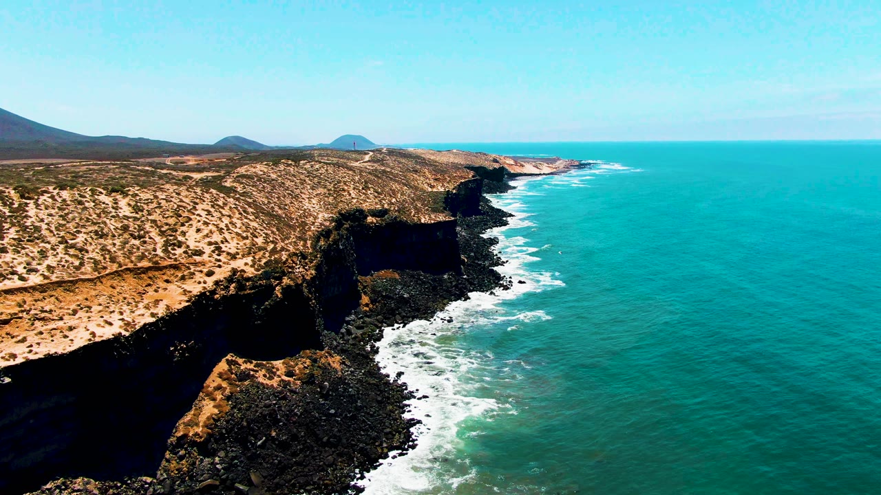 A slightly later view of the San Quintin BC Mexico headland shows white waves curling along the dark rocky base, dry scrub-covered land, open turquoise water, and no visible people.