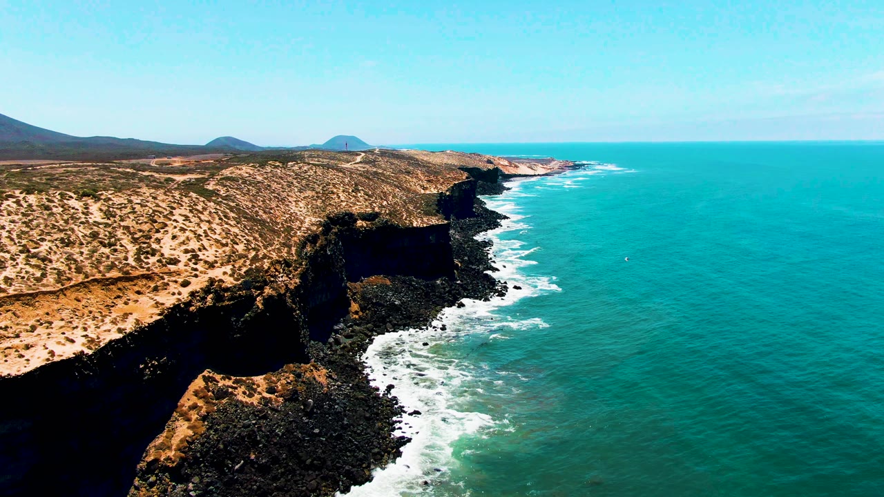The San Quintin BC Mexico coast fills the frame with tan bluffs on the left, a dark cliff pocket near center, turquoise ocean to the right, foamy surf below, and no visible people.