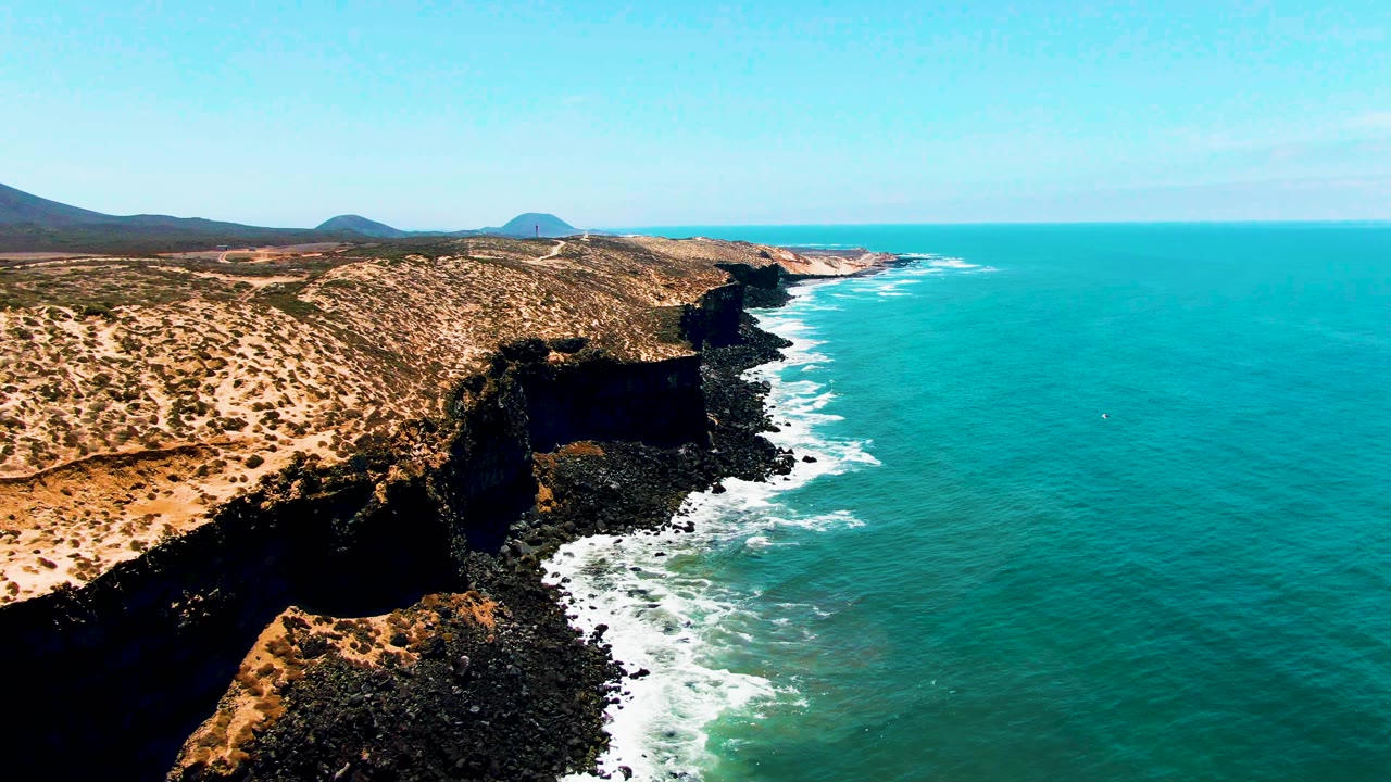 Midway through the clip, the San Quintin BC Mexico cliff ledge appears closer in the lower frame, with black rocks, white foam, scrubby arid ground, blue sky, and no visible people.