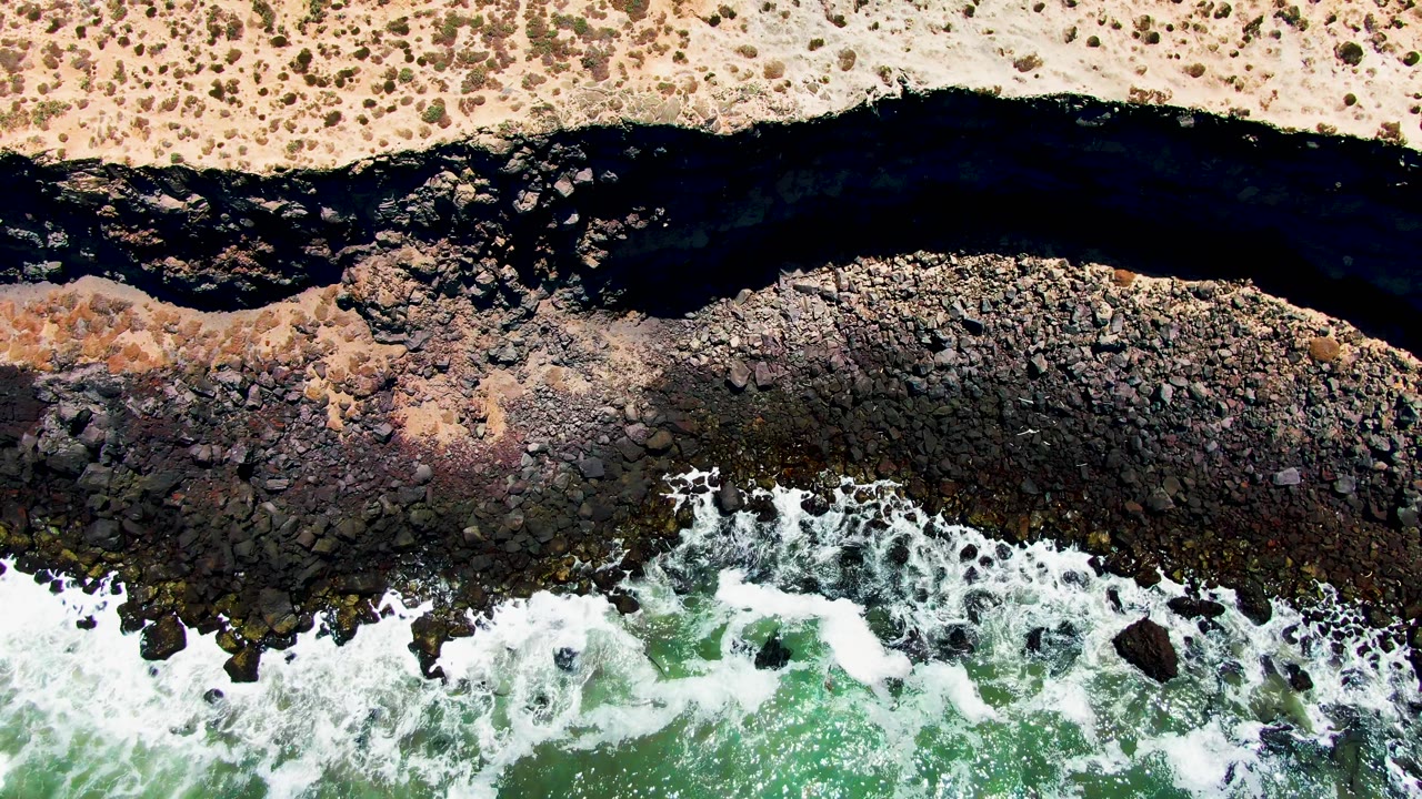 At 0:00, an overhead view in San Quintin BC Mexico shows sandy scrubland above a dark river channel, rocky shore, green surf, and no visible people.