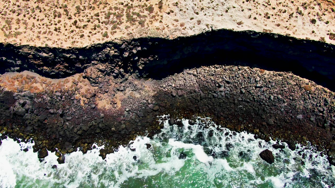 At 0:01, the San Quintin BC Mexico shoreline remains framed top-down, with white foam pushing across rocks below the dark river outlet and no visible people.