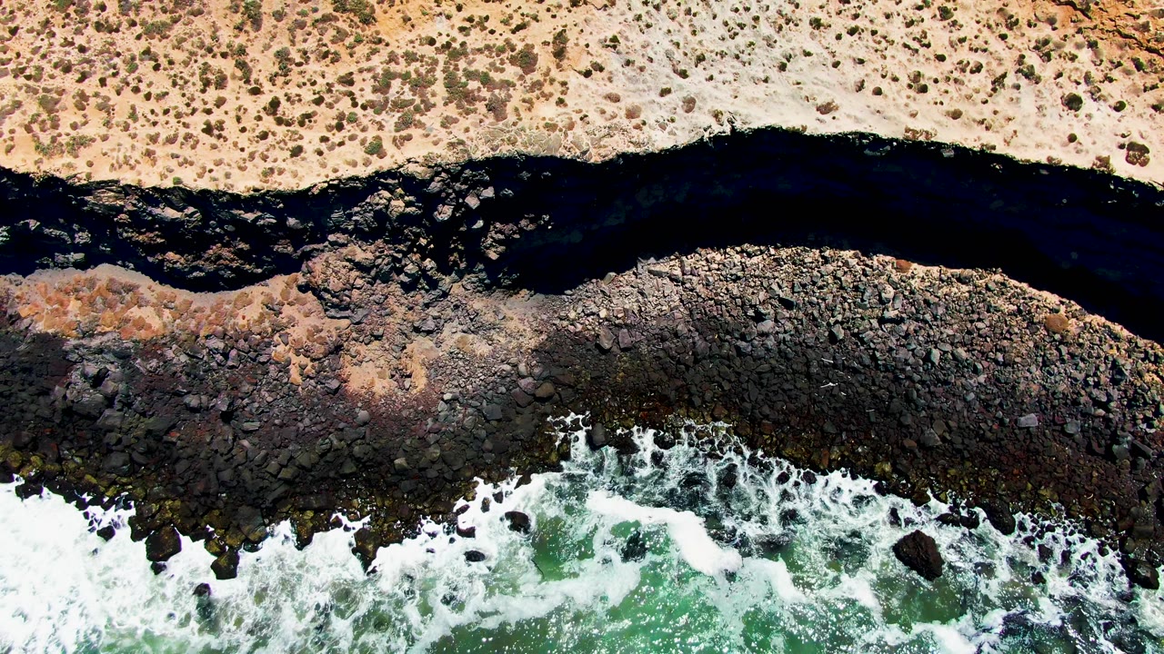 At 0:02, the aerial frame shows pale desert sand at the top, a winding dark channel, uneven coastal rocks, green water, white foam, and no visible people.