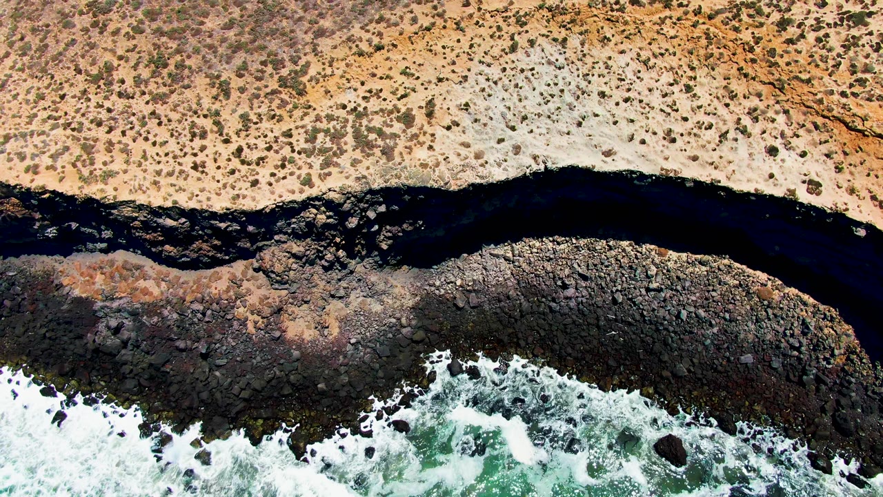 Overhead view in San Quintin BC Mexico showing tan scrubland, a dark river channel, rocky shoreline, white surf, and no visible people at the start of the clip.